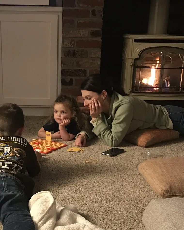 Dr. Kaci Kuykendall lying on the living room floor by the fireplace playing a board game with Baron, Ellis, and Blythe