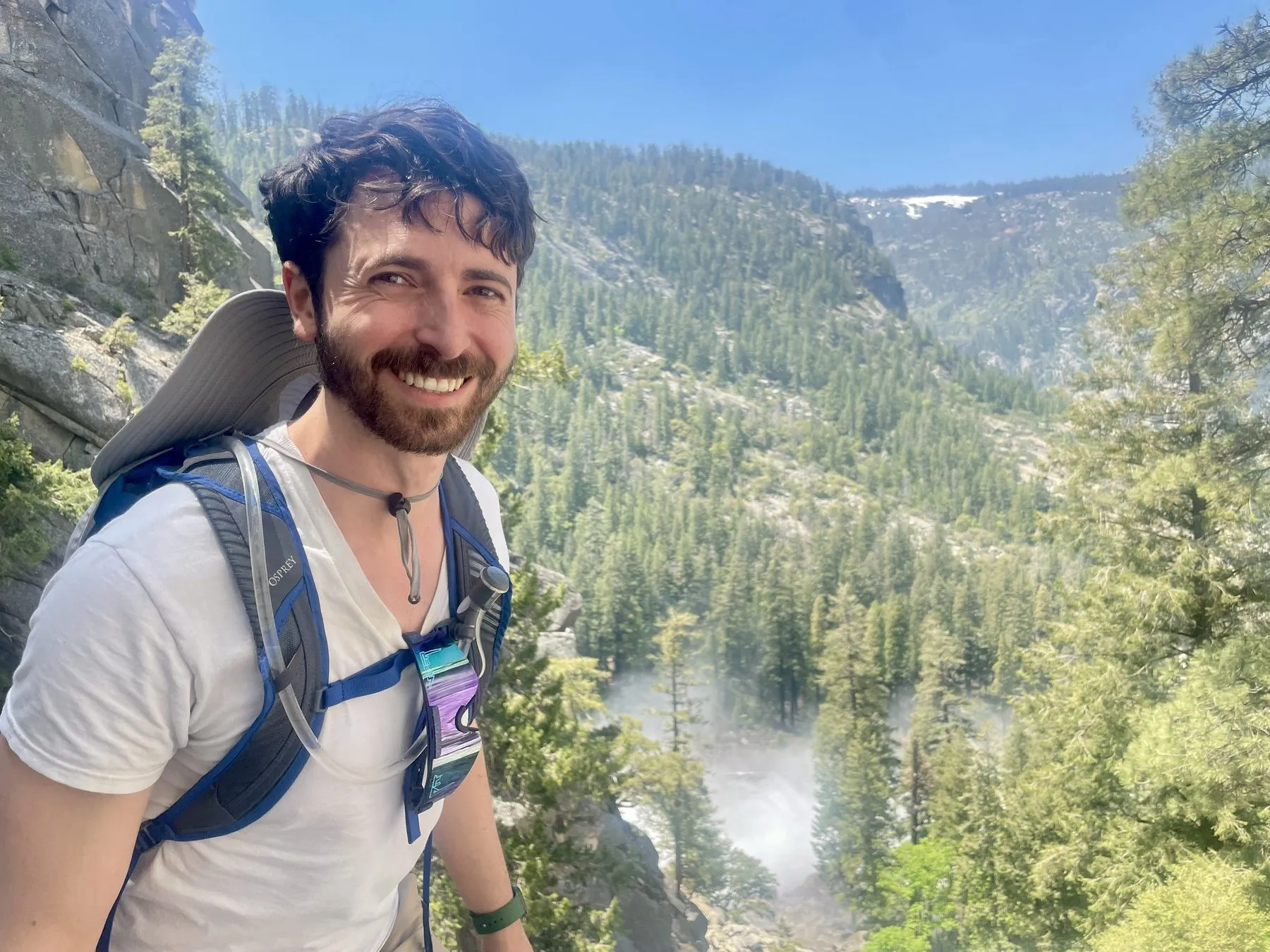 Dr. Rigel Smart on a mountain hiking trail wearing a backpack, with a forested canyon and waterfall behind him