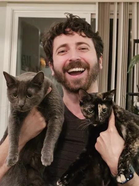 Dr. Rigel Smart laughing as he holds his two cats at home, with houseplants behind him